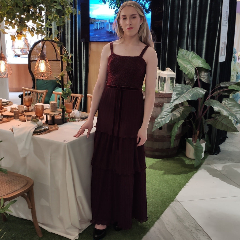 Ageless.ee deep burgundy tiered pleated maxi dress with embellished bodice, velvet waistband, and delicate straps. Model stands by a table setting.