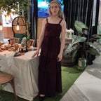 Ageless.ee deep burgundy tiered pleated maxi dress with embellished bodice, velvet waistband, and delicate straps. Model stands by a table setting.