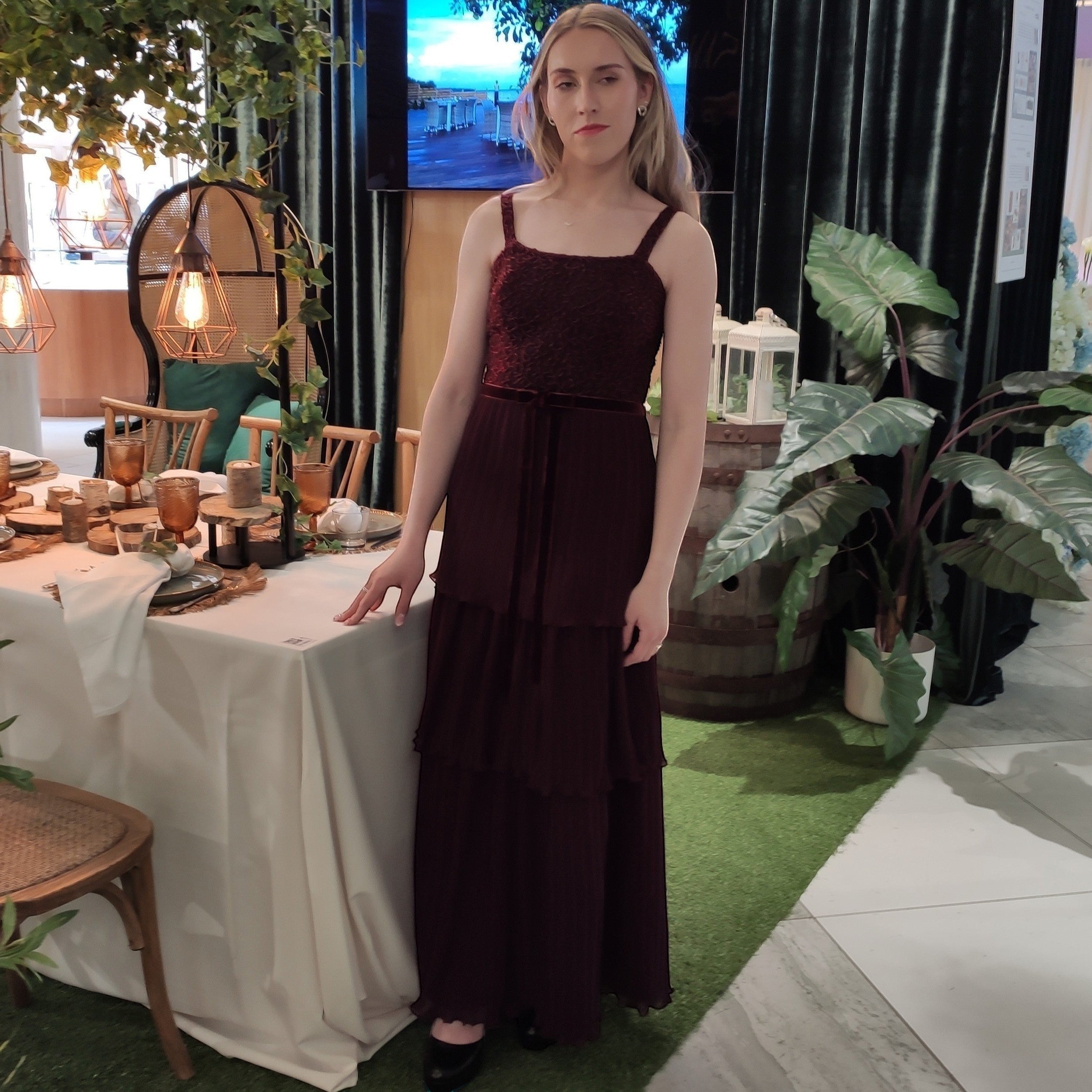 Ageless.ee deep burgundy tiered pleated maxi dress with embellished bodice, velvet waistband, and delicate straps. Model stands by a table setting.
