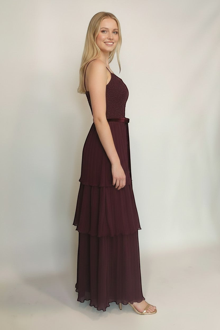 Ageless.ee burgundy tiered pleated maxi dress with embellished bodice, velvet waistband, and delicate straps. Model is wearing gold heels.